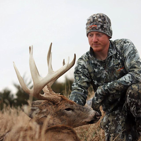 Hunter kneeling beside a large-antlered buck in grass, wearing First Lite camo and beanie