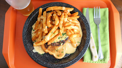 Venison sandwich topped with cheese sauce and fries on black plate, fork on green napkin