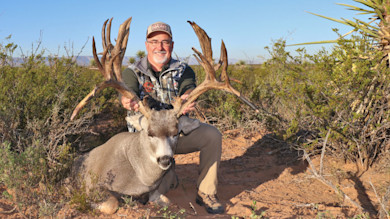 Hunter kneeling behind large deer with massive antlers in scrubby desert landscape