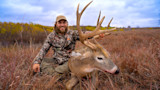Hunter in camouflage sitting in tall grass holding a large-antlered whitetail buck, smiling