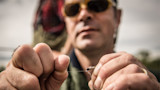 Hands tying a small fishing fly on a line, person blurred wearing sunglasses behind