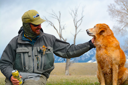 Angler in waders petting golden retriever with snow-capped mountains behind