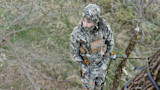 Hunter in full camouflage and face mask, wearing chest pack, secured to tree by climbing rope
