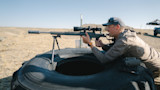 Man aiming scoped rifle on bipod over large tire at outdoor shooting range