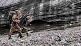 Two hunters with backpacks and rifles walking across rocky moraine below layered ice wall