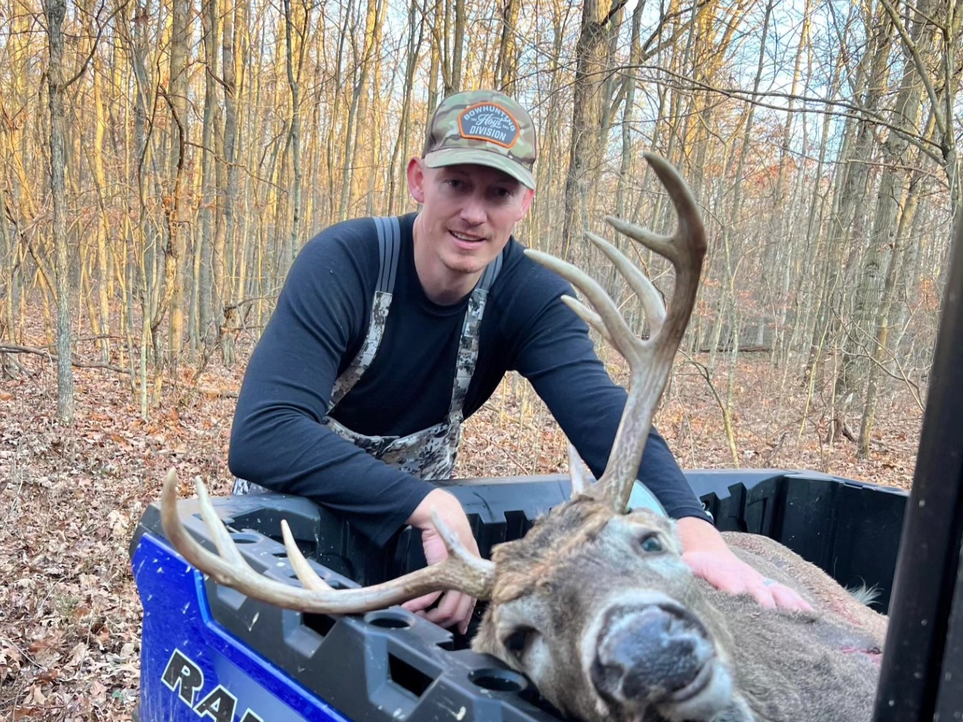 Hunter kneeling over buck with large antlers in an ATV bed in bare woods