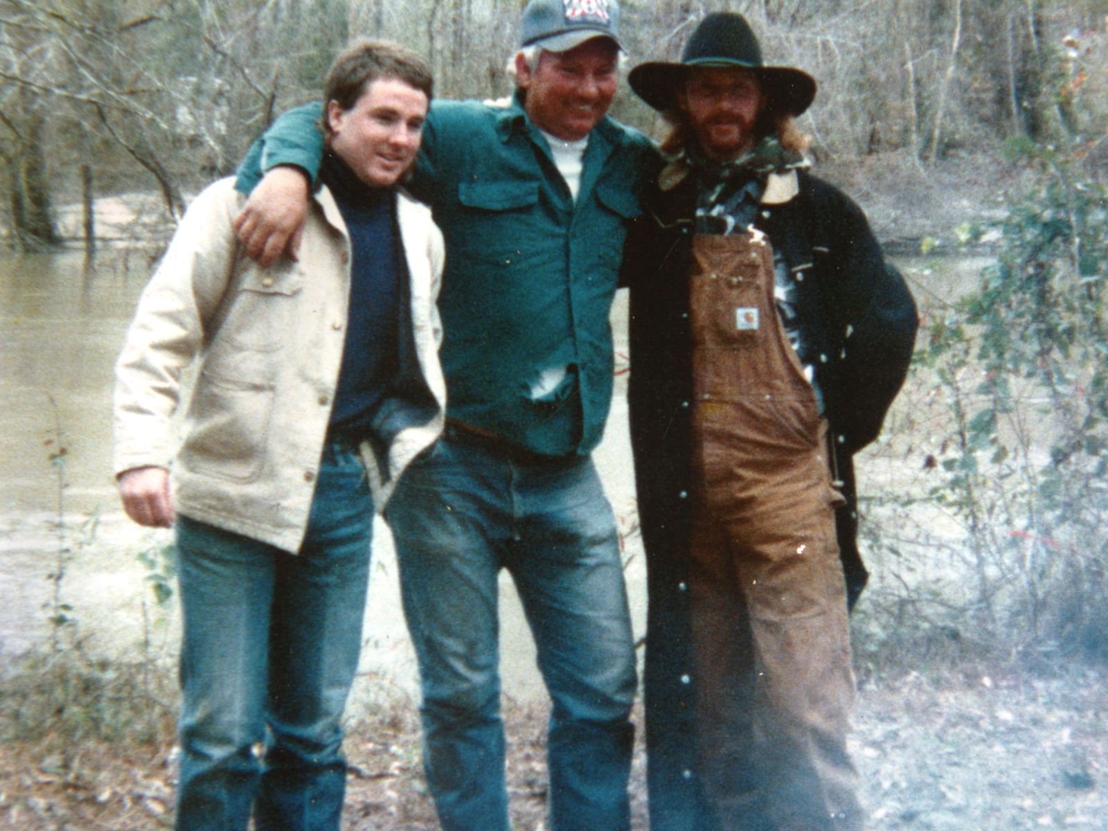 Three men arm-in-arm beside a flooded river in leafless woods