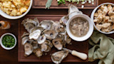 Shucked oysters on wooden board with bowl of oyster meat, bread, chopped herbs and a shucking knife