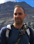 Man with backpack and sunglasses standing in front of steep rocky mountain cliffs