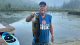 Man holding large smallmouth bass by a dock, wearing blue "Labatt Blue" sleeveless shirt.