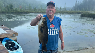 Man holding large smallmouth bass by a dock, wearing blue "Labatt Blue" sleeveless shirt.