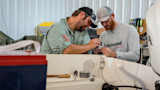 Two men inspecting wiring inside a small boat, one wearing a shirt reading "Lakewood"
