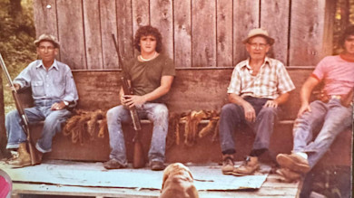 Four hunters seated on wooden bench by barn, two holding rifles, squirrel pelts between them, dog in foreground