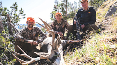 Three hunters kneel on a rocky hillside around a harvested mule deer buck; one holds a rifle.