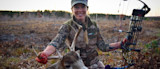 Woman hunter kneeling with buck and compound bow; vest reads 'OutfitHer'
