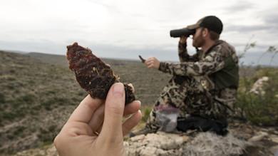 Hand holding jerky strip in foreground; hunter in camouflage using binoculars on ridge
