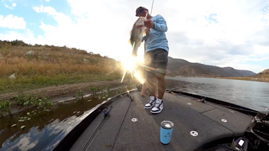 Angler holding large bass on boat deck with sun glare; fishing rods and blue tumbler visible