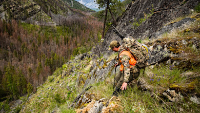 Hunter descending steep rocky slope wearing camouflage and orange vest, rifle on backpack