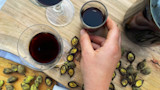 Hand holding small glass of dark walnut liqueur beside two wine glasses and cracked green walnut shells on wood