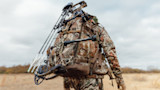 Hunter carrying camouflage hunting pack with compound bow and arrows in open field