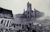 Ruined coastal town with damaged church, collapsed buildings and people amid debris.