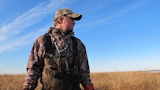 Hunter in camo waders and cap standing in marsh with duck calls around his neck