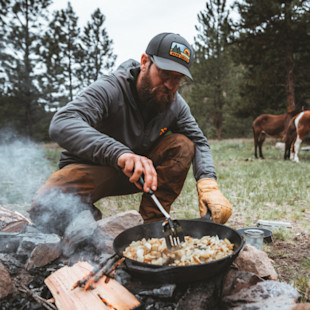 Clay Newcomb kneeling by a campfire, stirring diced potatoes in a cast-iron pan