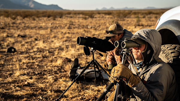 Two hunters using binoculars and a spotting scope on tripods beside a truck in dry plains