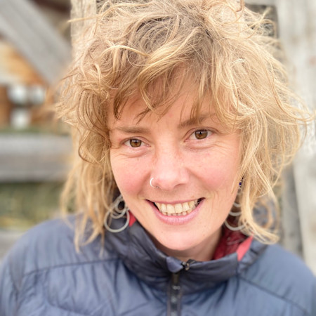 Smiling woman with curly blonde hair, nose ring and hoop earrings, wearing a gray puffer jacket