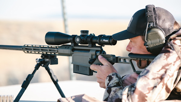 Man aiming rifle on bipod with SIG Sauer scope, wearing camo and earmuffs