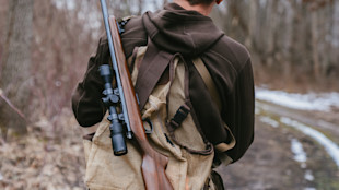 Hunter carrying scoped rifle slung over mesh backpack on muddy trail with patches of snow