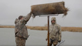 Two hunters on a boat mounting a rolled hay bale onto a raised feeder in a marsh