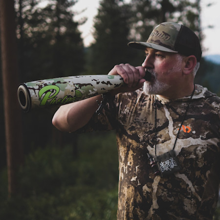 Hunter blowing a camo Phelps call in forest, wearing camouflage hoodie with orange chest logo