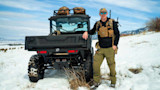 Man in sunglasses and chest rig leaning on ATV with rifle on pack in snowy field