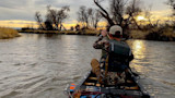 Hunter paddling a canoe at sunset on a river; camo jacket, life vest, bow and arrows with orange vanes
