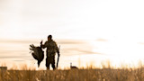 Hunter holding a wild turkey by the feet and a shotgun in a grassy field at sunrise