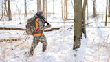 Hunter carrying rifle over shoulder and backpack through snow-covered woods, wearing camo and orange vest