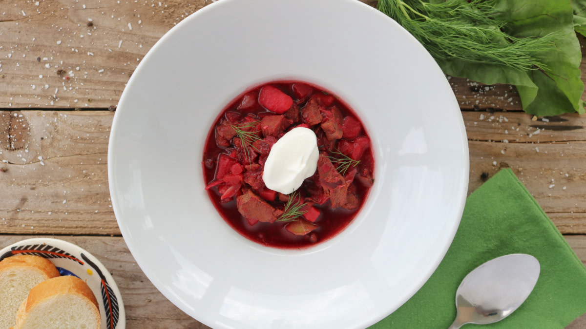 Borscht in white bowl topped with sour cream and dill; bread and spoon on wood