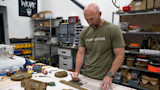 Man in workshop measuring webbing on table, wearing shirt reading "FHF GEAR"