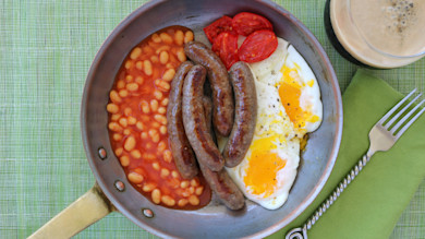 Skillet with five Irish sausages, baked beans, two fried eggs and roasted tomato; coffee cup and fork