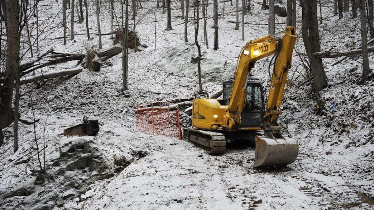 Yellow excavator parked on snowy hillside in leafless woods beside orange safety fence