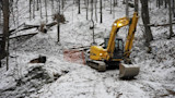 Yellow excavator parked on snowy hillside in leafless woods beside orange safety fence