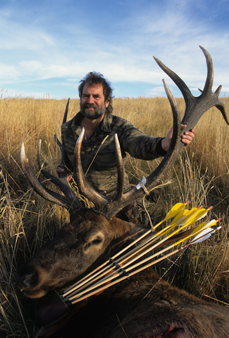 Hunter kneeling in tall grass holding large elk antlers beside elk with bow and yellow-fletched arrows