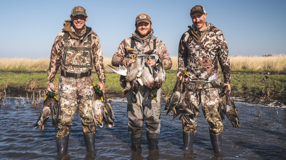 Three hunters in camo waders standing in shallow water holding harvested ducks; SIMMS logo on chest