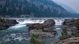 Wide river waterfall cascading over rocky ledges with forested mountains in background