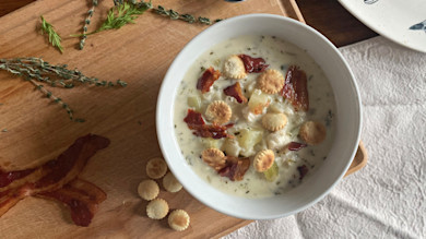 Creamy fish chowder in white bowl topped with oyster crackers, bacon and potatoes on wooden board