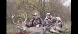Two hunters kneeling beside a large buck with antlers and a compound bow in woods