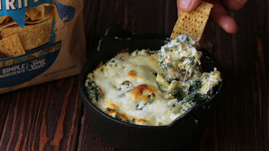 Cheesy stinging nettle and artichoke dip in a black skillet, cracker scooping; chip bag reads SIMPLE INGREDIENTS
