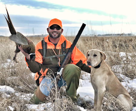 Hunter kneeling in snowy marsh holding pheasant and shotgun, labrador beside him, hat reads Cabelas