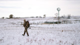 Hunter in camouflage carrying a compound bow and red-fletched arrows across a snowy field by a windmill and corral